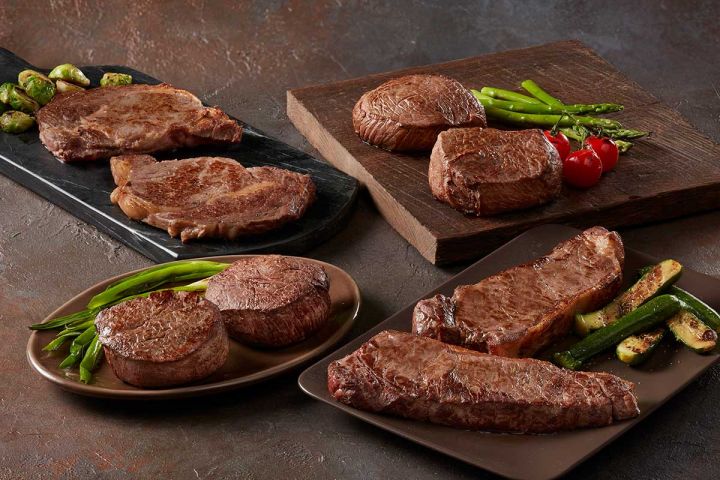 Assorted steaks and vegetables on plates and a cutting board with a dark background