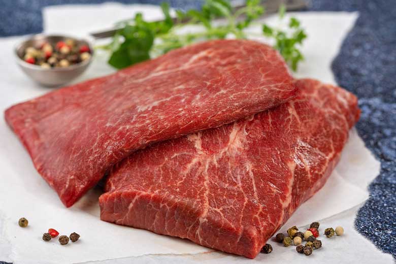 Two pieces of raw red meat on a white surface with peppercorns and herbs.